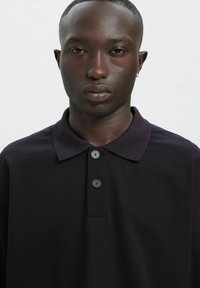 Czarna koszulka polo z teksturowanej tkaniny, z kołnierzem i plisą zapinaną na dwa guziki, prezentowana na jednolitym białym tle.