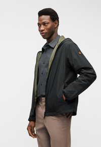 Άνδρας που φοράει μαύρο μπουφάν με κουκούλα πάνω από σκούρο γκρι πουκάμισο με κουμπιά και μπεζ παντελόνι, στέκεται με το ένα χέρι στην τσέπη του μπουφάν.