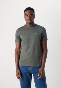 Tricou scurt cu mânecă, de culoare verde măslinie, dintr-un amestec de bumbac, având guler rotund și un mic logo alb pe piept, asortat cu blugi din denim albastru.
