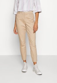 Pantalones de algodón beige con un corte ajustado, de longitud recortada y bolsillos frontales. Combinados con zapatillas blancas de caña alta y una camisa a rayas.