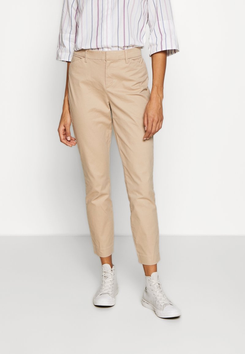 Pantalones de algodón beige con un corte ajustado, de longitud recortada y bolsillos frontales. Combinados con zapatillas blancas de caña alta y una camisa a rayas.