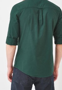 Zielona koszula z guzikami, z podwiniętymi rękawami, z plisą z tyłu i zaokrąglonym dolnym brzegiem, wykonana z gładkiego, lekkiego materiału.