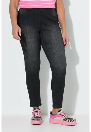 Frau trägt schwarze Skinny-Jeans und rosa Sneakers mit Leopardenmuster, steht vor einem weißen Hintergrund.