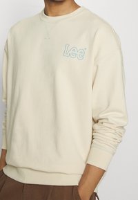 Krémová mikina vyrobená z měkké látky. Má světle modrou vyšitou značku "Lee" na hrudi a žebrované manžety a lem.