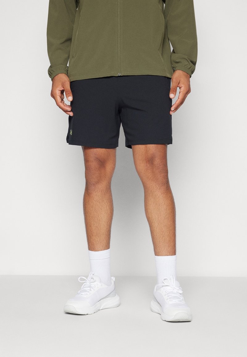 Pantalones cortos negros de corte ajustado, fabricados con tela ligera. Combinados con zapatillas blancas y una chaqueta con cremallera de color verde oliva.