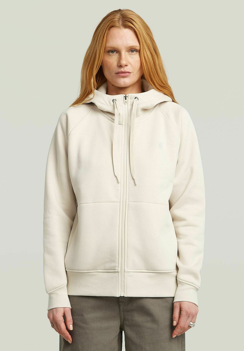 Woman with straight red hair wearing a cream zip-up hoodie with drawstrings and gray pants, facing forward against a plain background.
