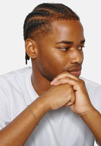 Giovane uomo con capelli intrecciati, che indossa una maglietta bianca e un bracciale d'oro, appoggiando il mento su mani unite su uno sfondo bianco.