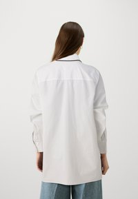 Vit oversized skjorta med en svart kant runt kragen, långa ärmar och rak skärning, tillverkad av lättviktig tyg.