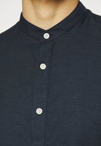Camicia button-up blu scuro con colletto a stand, caratterizzata da un motivo testurizzato e due bottoni bianchi sulla patta frontale.