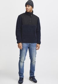 Navy fleece pullover with black panel and half-zip, worn with light blue jeans and black athletic shoes, topped with a gray knit beanie.