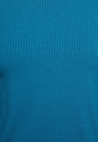 Rippstricktop in einem lebhaften Türkis. Es hat eine glatte Textur mit vertikalen Streifen, die das figurbetonte Design unterstreichen.