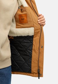 Brūns ārējais mētelis ar fliša oderi un melnu quiltu iekšpusi. Ietver uzšuvumu ar tekstu un kabatu ar rāvējslēdzēju.