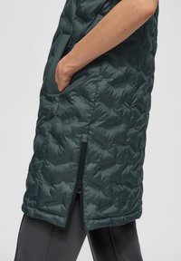 Grüner gepolsterter Mantel mit glänzender Oberfläche, seitlichen Taschen und einem Reißverschluss am unteren Saum, mit einem gesteppten Muster zur Isolierung.