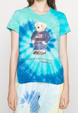 Persoon draagt een blauwgroen tie-dye T-shirt met een beer in spijkerbroek en een trui met de Amerikaanse vlag met tekst "Polo Bear by Ralph Lauren".