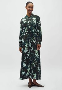 Femme souriante portant une robe maxi à manches longues boutonnée avec un motif floral abstrait en vert foncé, bleu et blanc, et des ballerines marron à bouts pointus.