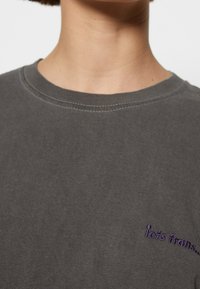 Gros plan sur le cou et le haut de la poitrine d'une personne portant un t-shirt gris foncé à col rond avec un petit texte brodé violet sur le côté droit.