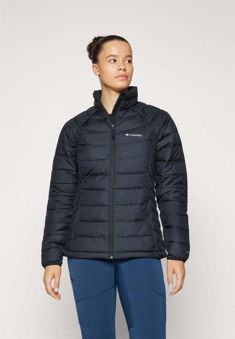 Giacca a compressione nera con colletto alto, design trapuntato, cerniera frontale e un logo sul petto sinistro. Indossata sopra pantaloni blu.
