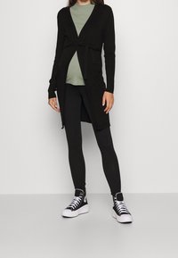 Černý kardigan se zavazováním v pase, nošený přes světle zelený top. Kombinován s černými legínami a vysokými černými teniskami. Jednoduchý, elegantní design.