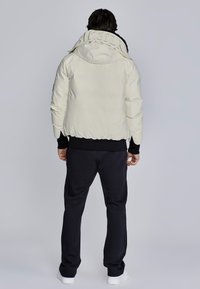 Giacca bianca imbottita con colletto alto e dettaglio logo sul collo, abbinata a pantaloni sportivi scuri e sneakers bianche. Vista posteriore.