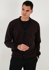 Cardigan color bordeaux scuro con scollo a V, chiusura anteriore con bottoni neri e polsini a costine. Realizzato in un tessuto morbido, presenta una vestibilità comoda e una trama uniforme.