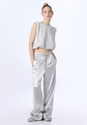 Giovane donna che indossa un top corto grigio senza maniche con un motivo sottile e pantaloni larghi in raso argentato, in piedi con le mani nelle tasche.