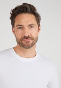 Chemise blanche à manches courtes avec un col rond, présentant une texture lisse et un design minimaliste, portée par un homme avec une barbe et des cheveux clairs.