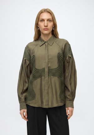 Femme portant un chemisier vert olive à manches longues avec des motifs ondulés texturés et un pantalon noir, debout devant un fond clair uni.