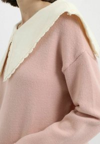 Mujer vistiendo un suéter de punto rosa pálido con un amplio cuello color crema que presenta un borde ondulado.