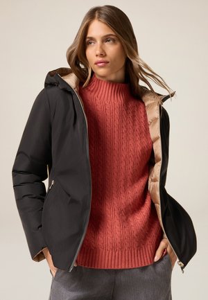 Chaqueta acolchada negra con cierre frontal, con un forro interior marrón. Llevada sobre un suéter rojo texturizado. Fondo neutral y claro.