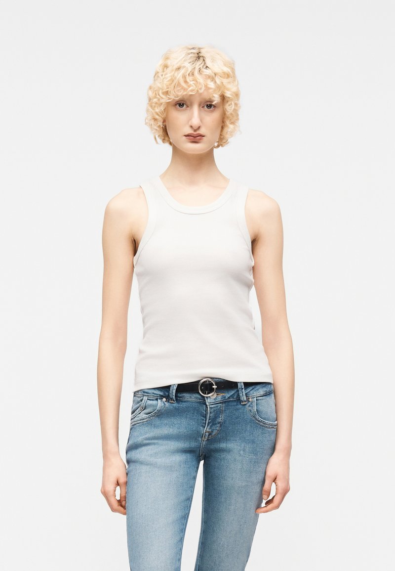 Personne blonde aux cheveux bouclés portant un haut blanc sans manches et un jean bleu clair avec une ceinture noire, debout devant un fond uni.