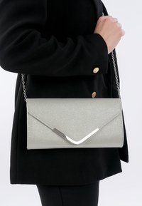 Persoon met zwarte jas houdt een zilverkleurige clutch met envelopstijl en kettingriem vast tegen een witte achtergrond.