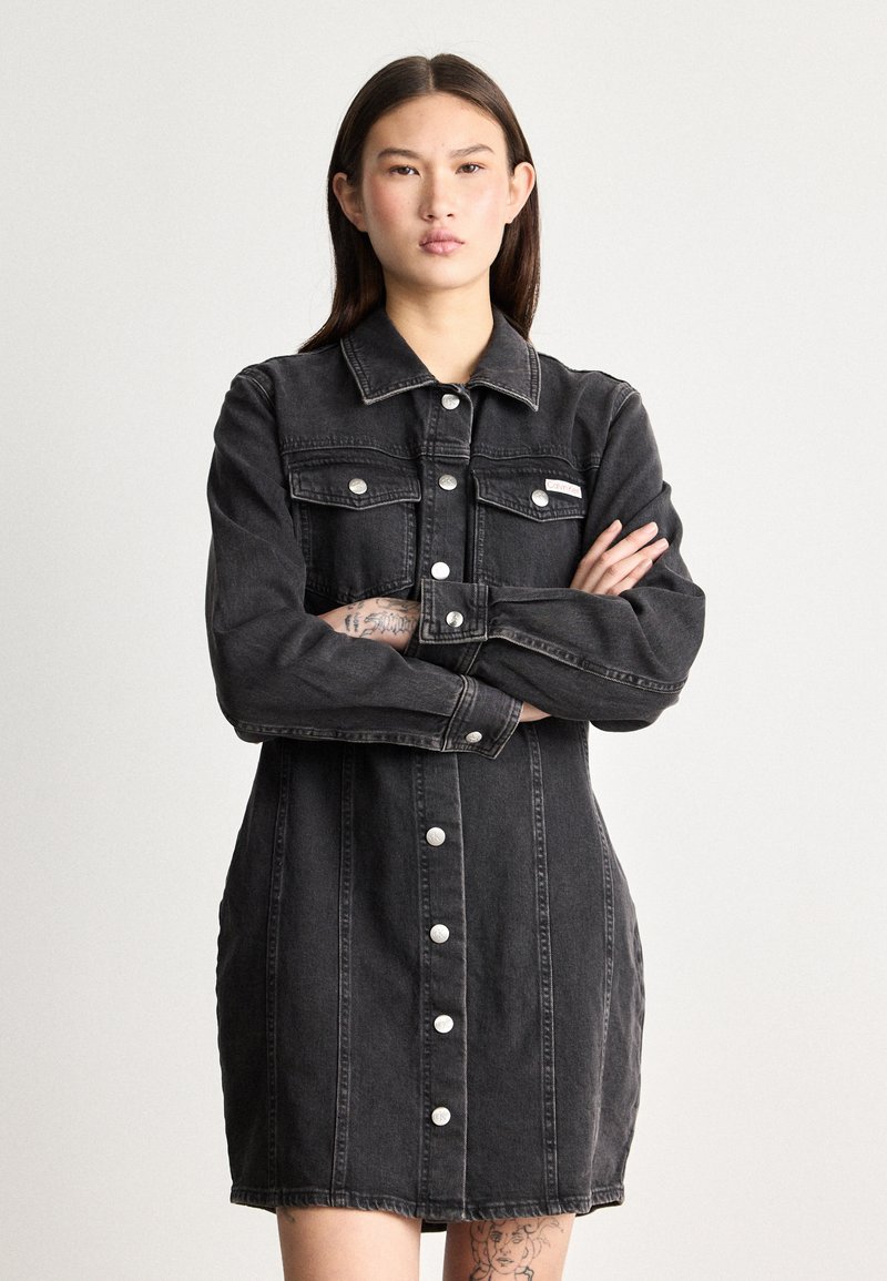 Vestido de ganga preta com fecho de botões, gola pontuda, dois bolsos no peito e mangas longas. Apresenta uma bainha desfeita e detalhes de costura subtis.