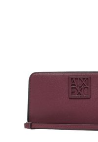 Portafoglio in pelle bordeaux con zip a tutto giro, dotato di placca con logo Armani Exchange e tracolla attaccata su sfondo bianco.