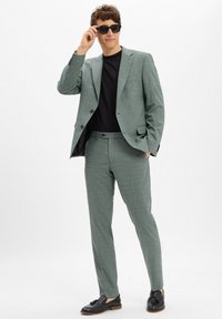 Hellgrüner Karosakk mit taillierter Passform, schwarzes T-Shirt darunter und schwarze Schuhe. Mann, der sich die Sonnenbrille anpasst, leichter Hintergrund.