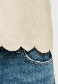 Haut en tissu beige avec ourlet festonné, porté sur un jean en denim bleu avec une poche avant et des coutures apparentes.