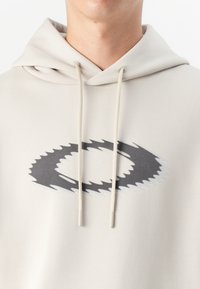 Smėlinis džemperis su apvaliu, dantytu juodu logotipu priekyje, su raišteliais ir minkštu audinio tekstūru.