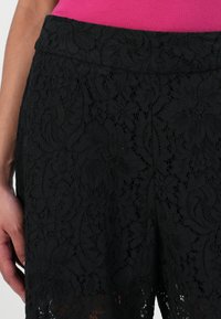 Shorts en dentelle noire avec un motif floral, dotés d'une taille lisse et d'une texture ajourée, révélant la peau en dessous.