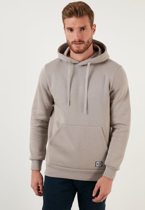 Sudadera gris claro hecha de tejido suave. Cuenta con un bolsillo tipo canguro, cordones, puños acanalados y un pequeño parche con el logo en el dobladillo.