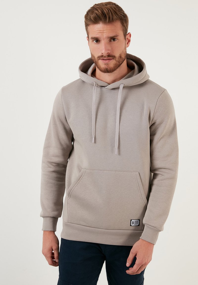 Sudadera gris claro hecha de tejido suave. Cuenta con un bolsillo tipo canguro, cordones, puños acanalados y un pequeño parche con el logo en el dobladillo.