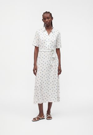 Femme portant une robe midi blanche avec de petits imprimés floraux bleus, des manches courtes, une ceinture nouée à la taille, et des sandales blanches plates à lanières.
