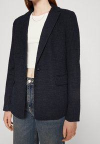 Námořnický texturovaný blazer s jedním knoflíkem, klopovým límcem a kapsami na nášivkách, nošený přes bílý crop top a spárovaný s džíny.