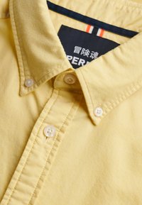 Chemise jaune à boutons avec un col boutonné et une étiquette noire visible comportant un texte blanc et rouge ainsi que des caractères japonais.