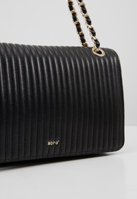 Sac à main en cuir noir matelassé avec coutures verticales, bandoulière en chaîne dorée entrelacée de cuir noir, et petit logo doré "abro" à l'avant.