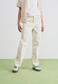 Pantalone boje kreme, slobodnog kroja od lagane tkanine sa ravnim nogavicama. Upareni sa bijelom košuljom i cipelama s zelenim akcentima.