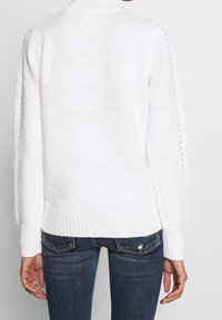 Vit stickad tröja med hög krage, texturerat mönster och ribbade muddarna. Baksidan har en lös passform och subtila sidodetaljer.