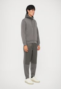 Felpa grigia con zip e cappuccio decorato, abbinata a pantaloni jogger grigi con pannelli a contrasto; scarpe da ginnastica bianche completano l'outfit.