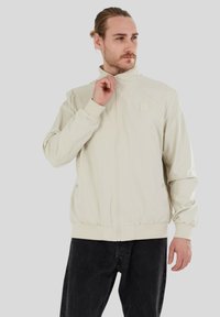 Helle beige Bomberjacke mit einem Reißverschluss vorne, elastischen Bündchen und Saum. Verfügt über ein kleines Logo-Patch auf der Brust und eine glatte Textur.
