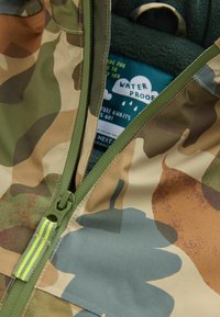 Jakke med camouflagemønster i jordfarver; har en vandtæt ydre, grøn lynlås og en blød foret inderside med en sky-grafiklabel.