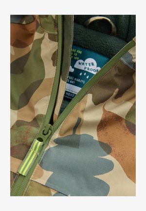 Veste à motifs camouflage dans des tons terreux ; présente un extérieur imperméable, une fermeture éclair verte et une doublure intérieure douce avec une étiquette graphique en forme de nuage.