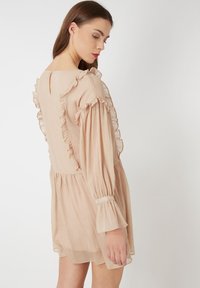 Vestido de manga corta, de color beige claro, con detalles de volantes en las mangas y la parte trasera. Fabricado con un tejido ligero y texturizado. Presenta un escote en la espalda.